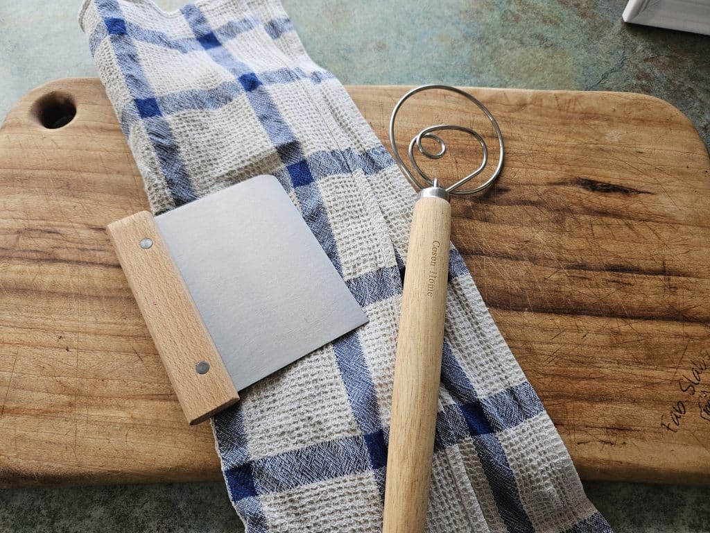 Dough whisk and scraper on a cutting board.