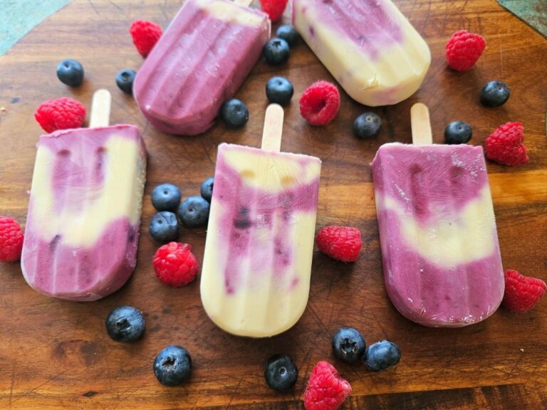 Berry cheesecake popsicles on a cutting board surrounded by berries.