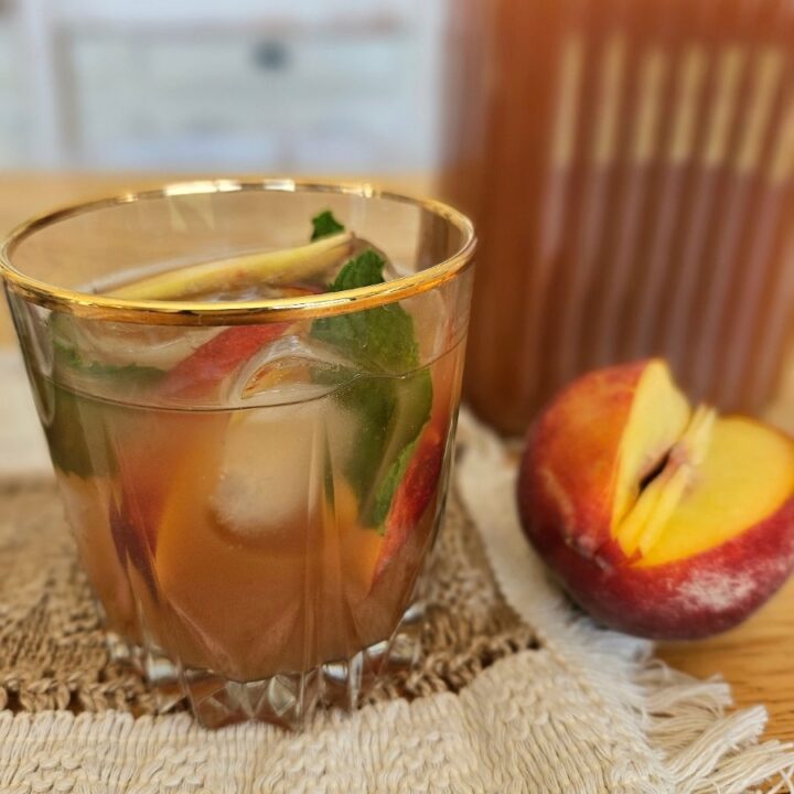 Beautiful glass filled with peach iced tea, peppermint leaves and slice peaches.