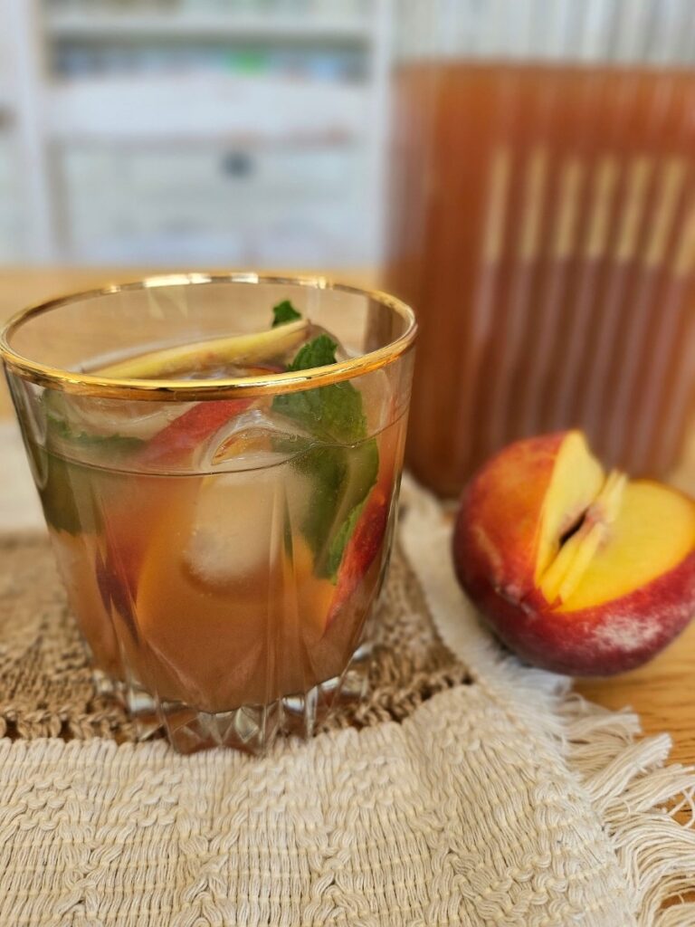 Beautiful glass filled with peach iced tea, peppermint leaves and slice peaches.