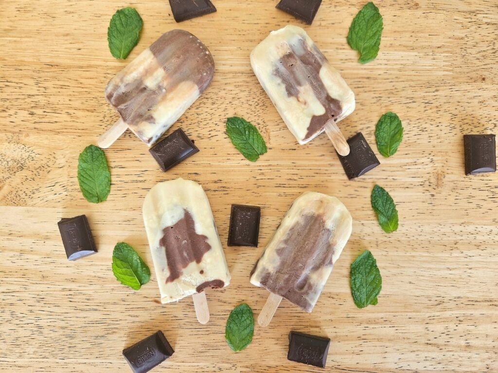 Homemade creamy mint chocolate popsicles on a table surrounded by chocolate and mint.