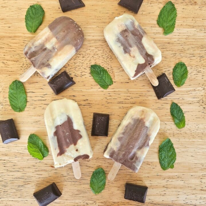 Homemade creamy mint chocolate popsicles on a table surrounded by chocolate and mint.