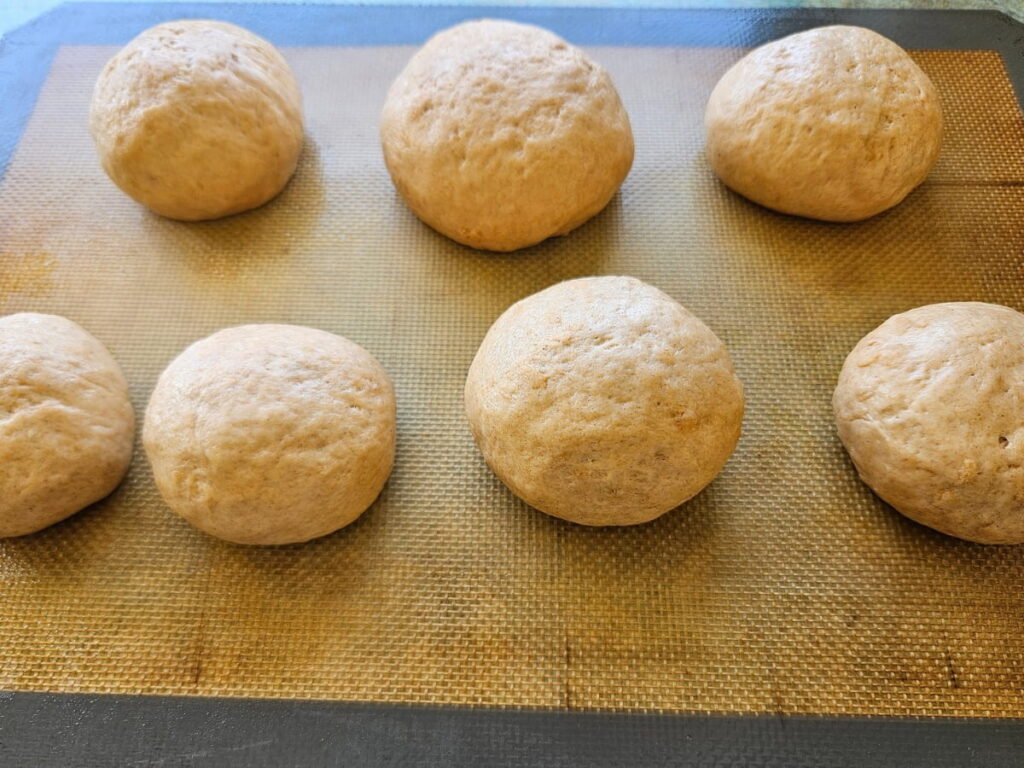 Hamburger buns rising on a silicone mat on kitchen counter.